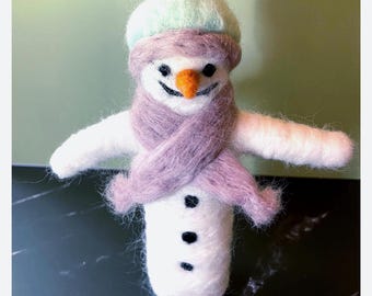 Könnte beinhalten: Eine aus weißer Wolle gefilzte Schneemannfigur mit einer hellgrünen Bommelmütze, einem weichen lavendelfarbenen Schal, einer leuchtend orangefarbenen Karottennase, schwarzen Perlenaugen, einem gestickten Lächeln und drei kleinen schwarzen Knöpfen.