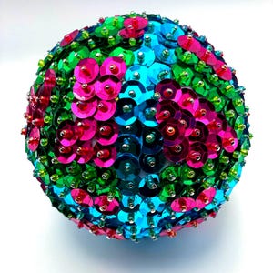 May include: A spherical ornament covered in colorful sequins. The sequins are in shades of pink, green, and blue, creating a vibrant, textured surface. The ornament is likely for holiday or decorative use.