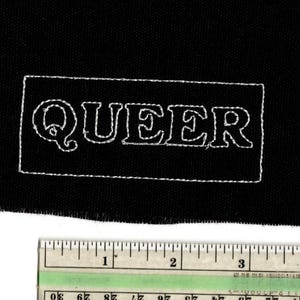 May include: A black fabric patch with the word "QUEER" embroidered in white. The text is inside a white rectangular border. A ruler is placed below the patch, showing measurements in inches.