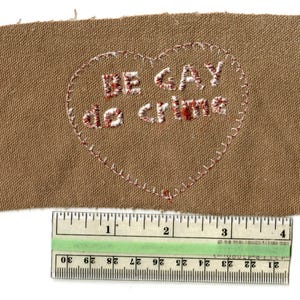 May include: A brown fabric patch with frayed edges, featuring embroidered text within a heart shape. The text reads "BE GAY do crime" in a mix of red and white thread. A ruler is placed below the patch, showing measurements in inches.