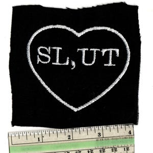 May include: Black embroidered patch with a white heart outline. The word "SLUT" is embroidered in white inside the heart. A ruler is at the bottom of the patch, showing inches.