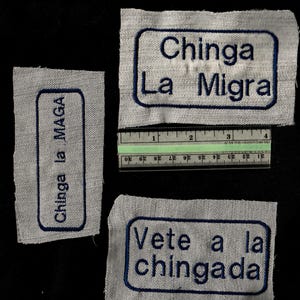 May include: Three embroidered patches with blue text on a light gray fabric. The patches feature the phrases "Chinga La Migra", "Chinga la MAGA", and "Vete a la chingada". A ruler is placed near the patches.