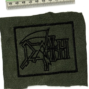May include: A dark green fabric patch with a black embroidered design. The design features a stylized scythe and the letters "DACH". A ruler is placed above the patch, showing measurements in inches.