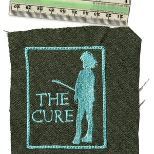 May include: Embroidered patch featuring the band name "THE CURE" and a teal silhouette of a person holding a guitar. The patch is on a dark green fabric, with a teal border. A ruler is placed above the patch.