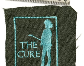May include: Embroidered patch featuring the band name "THE CURE" and a teal silhouette of a person holding a guitar. The patch is on a dark green fabric, with a teal border. A ruler is placed above the patch.