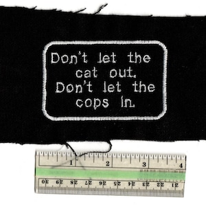May include: Black fabric patch with white embroidered text that reads "Don't let the cat out. Don't let the cops in." A ruler shows the patch is approximately 4 inches long. The patch has a rectangular shape with a white border.