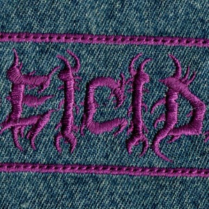 May include: A purple embroidered patch with the word "DEICIDE" in a gothic font, framed by a purple border. The patch is set against a blue denim background, showcasing a textured fabric.
