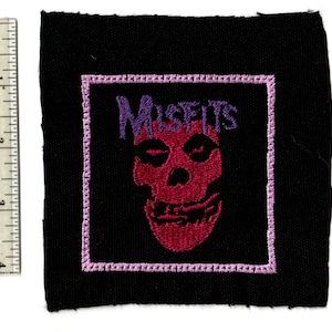 May include: Black square patch featuring a red skull with the word "MISFITS" in purple above it. The skull is framed by a light purple border. The patch is approximately 2 inches by 2 inches.