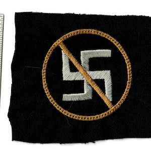 May include: Black fabric patch with a white swastika symbol inside a gold circle, crossed out with a gold diagonal line. The patch is approximately 2 inches wide.