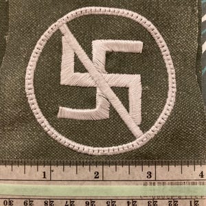 May include: Embroidered patch featuring a white swastika symbol inside a white circle with a diagonal line through it, indicating prohibition. The patch is on a green fabric background. A ruler is at the bottom.