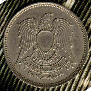 May include: A silver coin with the coat of arms of Syria. The coat of arms features an eagle with wings spread, a shield, and a wreath of laurel leaves. The coin also has Arabic text around the edge.