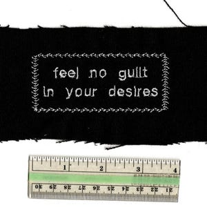 May include: Black fabric patch with white embroidered text: "feel no guilt in your desires." The text is framed by a decorative border. A ruler with inch measurements is at the bottom. The patch is approximately 3 inches wide.