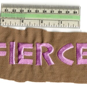 May include: A brown fabric patch with the word "FIERCE" embroidered in purple thread. A ruler is placed above the patch, showing measurements in inches. The patch is approximately 3 inches wide.