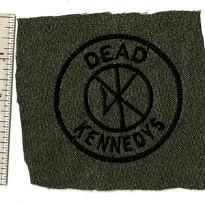 May include: A dark green fabric patch with the words "DEAD KENNEDYS" in black, encircling a black symbol. The patch is approximately 3 inches in length.