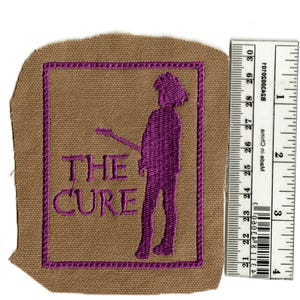 May include: Embroidered patch featuring the band name "THE CURE" in purple, along with a purple silhouette of a person playing a guitar. The patch is on a tan background with a purple border.
