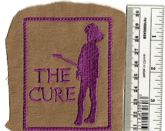 May include: Embroidered patch featuring the band name "THE CURE" in purple, along with a purple silhouette of a person playing a guitar. The patch is on a tan background with a purple border.