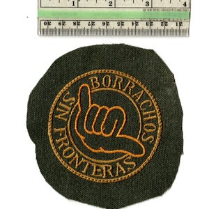 May include: A dark green patch with gold embroidered text and a hand gesture. The text reads "SIN FRONTERAS" and "BORRACHOS" in a circular design. A ruler is placed above the patch, showing measurements in inches.