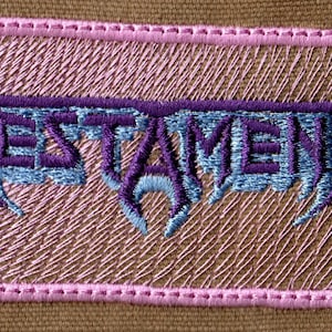 May include: Embroidered patch featuring the word "TESTAMENT" in a stylized, purple and light blue font. The patch has a pink border and is set against a tan background. The design is detailed and appears to be machine-embroidered.