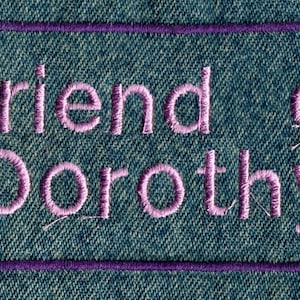 May include: A purple embroidered patch on a blue denim background with the text "Friend of Dorothy" in pink.