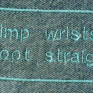 May include: A blue denim patch with white embroidered text that reads "limp wrists shoot straight".