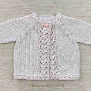 Könnte beinhalten: Ein hellrosa gestrickter Baby-Cardigan mit Knopfleiste und einem zarten Spitzenmuster auf der Vorderseite. Der Cardigan hat lange Ärmel und einen gerippten Saum. Der Text "Handknitsbyelena" ist am unteren Rand des Bildes sichtbar.