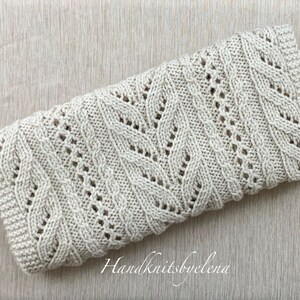 May include: An off-white knitted scarf with a cable and openwork pattern, folded neatly. The scarf is displayed on a light wooden surface. The text "Handknitsbyelena" is visible. The scarf's design is intricate.