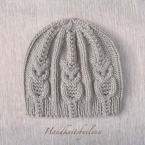 May include: A gray knitted beanie hat with a cable knit pattern and three small silver beads. The hat has a simple design and is made with a soft yarn.
