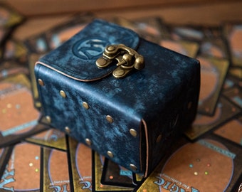 May include: A blue leather deck box with a brass clasp and rivets. The box is sitting on a pile of Magic: The Gathering cards.
