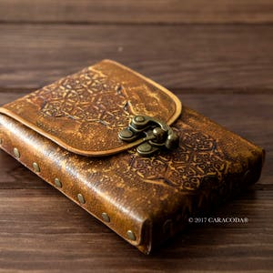 May include: A rectangular brown leather case featuring an embossed geometric pattern on the flap and body. The case is secured with a brass-toned metal latch and accented with small brass-toned rivets along the side. The text 2017 CARACODA is visible.