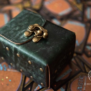 Peut inclure: Une boîte en cuir vert foncé avec un fermoir en laiton et des clous décoratifs. La boîte est posée sur une pile de cartes à jouer.