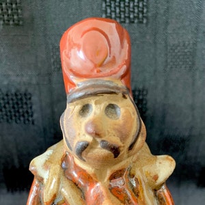 May include: A ceramic figurine of a soldier in a red coat and tall hat, holding a staff. The figurine has a mustache and is painted in shades of brown, red, and white. The figure is standing upright.
