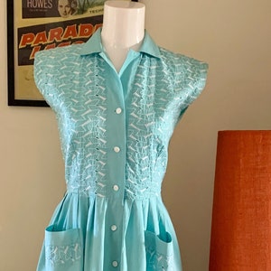 Peut inclure: Robe vintage bleu clair avec un col et des manches courtes. Le corsage présente un motif blanc et la robe a une fermeture boutonnée sur le devant et deux poches avant. La robe est présentée sur un mannequin.