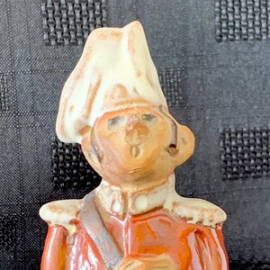 May include: A ceramic figurine of a soldier wearing a white hat, red jacket, and green pants. The soldier is holding a brown object in his right hand.