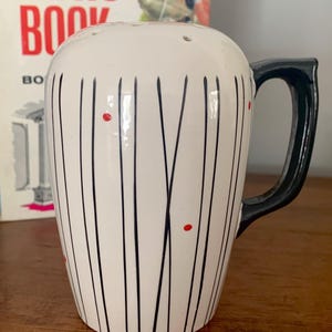 Può includere: Un dosatore in ceramica bianca con la parte superiore arrotondata dotata di molteplici fori. Il corpo è decorato con sottili linee verticali nere e piccoli punti rossi. Presenta un solido manico nero sul lato destro e una finitura lucida.