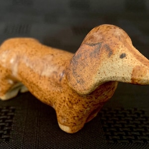 Pode incluir: Estatueta de dachshund em cerâmica, em tons de castanho e bege. O cão tem um corpo longo, pernas curtas e um rosto detalhado. A estatueta tem uma aparência rústica e texturizada, sugerindo que é um item decorativo.