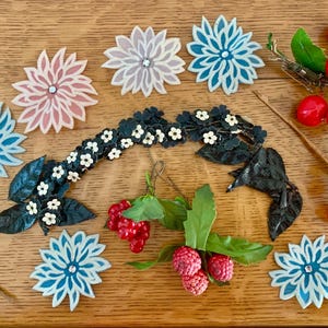Könnte beinhalten: Eine Auswahl an dekorativen Blumen- und Beerenverzierungen in verschiedenen Farben. Die Anordnung umfasst blaue, rosa und weiße Blumenformen sowie Büschel roter Beeren und grüne Blätter. Die Artikel sind auf einer Holzoberfläche angeordnet.