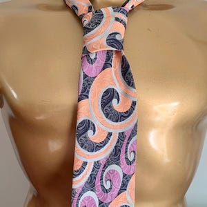 May include: A necktie featuring a repeating pattern of swirling shapes in shades of peach, lavender, and dark charcoal grey. The dark grey sections contain a fine, intricate paisley-like texture, while the peach and lavender swirls are solid.