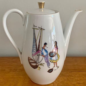 Pode incluir: Um bule de porcelana branca com um puxador de tampa dourado e uma alça curva. O corpo apresenta uma ilustração colorida de um veleiro, um pequeno barco a remos, uma pessoa junto ao barco e outra pessoa segurando uma cesta de peixes.