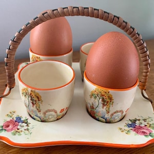 Puede incluir: Un juego de servir huevos vintage con cuatro hueveras y una bandeja. Las hueveras y la bandeja son de color crema con diseños florales y paisajísticos. La bandeja tiene un borde naranja y un asa trenzada.