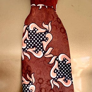 Può includere: Una cravatta rosso scuro caratterizzata da un motivo ripetitivo di quadrati a scacchi neri e bianchi accentuato da forme curve color pesca. La cravatta è annodata e poggia su un busto di manichino dorato con il testo Y-FRONT visibile in alto.