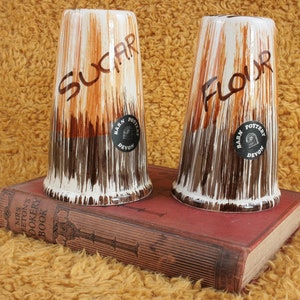 Op de afbeelding: Twee vintage keramische potten met een bruine en witte glazuur. De potten zijn gelabeld met "Suiker" en "Bloem" en hebben een zwarte sticker met de woorden "Barn Pottery Devon".