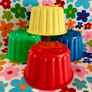 Könnte beinhalten: Ein Stapel von vier bunten, vintage Gelatineformen in Rot, Grün, Blau und Gelb. Jede Form hat ein geriffeltes Design und kleine Füße. Die Formen sind auf einem floral gemusterten Hintergrund angeordnet.