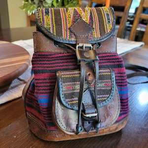 May include: A small backpack crafted from brown leather and woven fabric featuring horizontal stripes in red, black, and blue. The bag includes a top flap and a front pocket, both secured with leather straps, metal buckles, and decorative patterns.