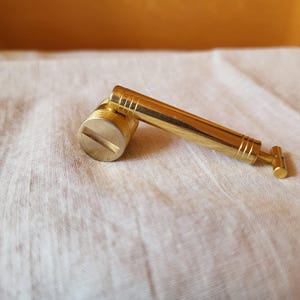 May include: A brass-colored, angled tool with a cylindrical head and a handle. The head has a slot for a screwdriver, and the handle has decorative ridges. The tool is on a white fabric surface.