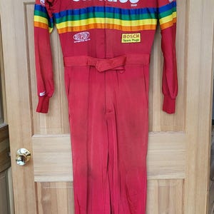 May include: A red racing suit with a rainbow stripe across the chest and Skittles branding. The suit has several sponsor logos, including NASCAR, Winston Cup Series, Pontiac, Valvoline, Starburst, Goodyear, Simpson, DuPont, and Bosch.