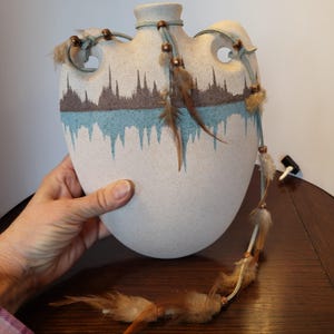 May include: A white ceramic vase with a brown and blue design and a fringe of feathers and beads hanging from the top.