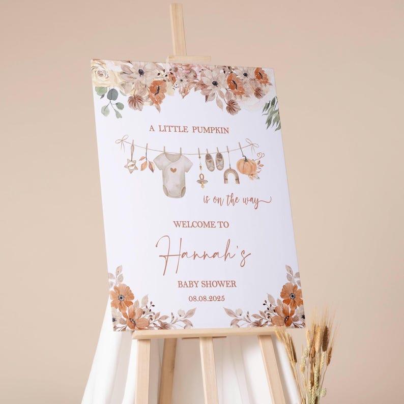 May include: A white baby shower sign on an easel, decorated with floral accents in shades of brown and orange. The sign reads "A Little Pumpkin is on the way" and "Welcome to Hannah's Baby Shower 08.08.2025".