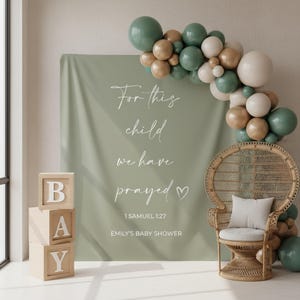 May include: Sage green backdrop with white script: "For this child we have prayed." Wooden blocks spell "BAY." A balloon arch in green, gold, and white accents the scene. A wicker peacock chair with a white cushion completes the baby shower decor.