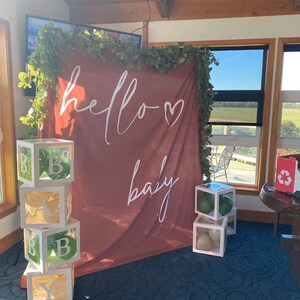 Puede incluir: Un tel&oacute;n de fondo rojo con texto blanco que dice "hello baby" con un coraz&oacute;n. Hay cajas transparentes apiladas una encima de la otra con letras y globos verdes y blancos en su interior.
