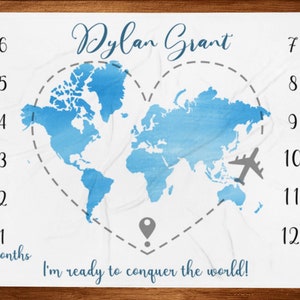 Puede incluir: Manta de hito blanca con un diseño de mapa del mundo en acuarela azul. La manta presenta el nombre "Dylan Grant" y la frase "I'm ready to conquer the world!" con los números 1-12 y un avión gris.